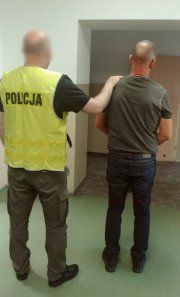 W korytarzu komisariatu na Żoliborzu policjant oczekuje z zatrzymanym mężczyzną na czynności procesowe. Policjant ubrany w kamizelkę z napisem POLICJA, trzyma rękę na plecach zatrzymanego. Obaj stoją tyłem do obiektywu.