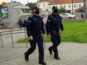 Na pierwszym planie widzimy dwóch policjantów w mundurach, którzy patrolują Warszawę pod kątem przestrzegania obostrzeń związanych z wprowadzeniem obowiązku noszenia maseczek w przestrzeni publicznej