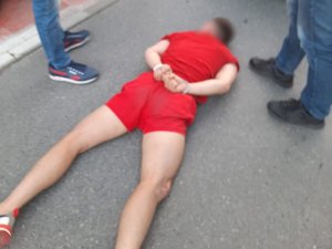 Zatrzymany mężczyzna leży na ziemi z założonymi kajdankami na ręce trzymane z tyłu. Obok niego stoją pilnujący go policjanci.