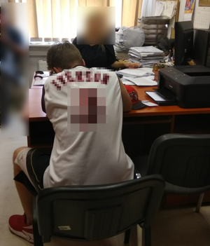 Zdjęcie przedstawia siedzącego tyłem mężczyznę w białej koszulce, zasłoniętym napisem na plecach. Naprzeciwko niego przy biurku siedzi policjantka, która przesłuchuje mężczyznę. Na biurku widać teczki z dokumentami.