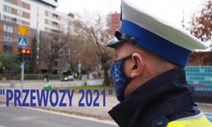 Zdjęcie przedstawia policjanta ruchu drogowego na tle skrzyżowania. Na zdjęciu widnieje napis PRZEWOZY 2021