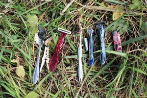 Zdjęcie przedstawia leżące na trawie przedmioty: metalowa łyżka, maszynki do golenia, zapalniczka, szczoteczka do zębów, spinacz do ubrań.
