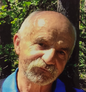 Zdjęcie przedstawia twarz mężczyzny w wieku około 70 lat, ma on krótkie siwe włosy, łysinę czołową, patrzy na wprost obiektywu aparatu. Widać fragment niebieskiej koszulki.