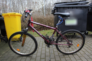zdjęcie przedstawia podparty na nóżce rower typu górskiego w kolorze bordowym, który stoi na dworze na tle żółtego i czarnego kontenera na śmieci.