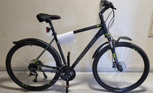 Zdjęcie przedstawia rower stojący w pomieszczeniu. Rower jest ciemny, posiada zielone elementy. Na ramie ma zawieszoną białą kartkę.