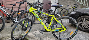 Zdjęcie przedstawia rower w kolorze żółtym z ciemnymi kołami stojący na tle innych rowerów.
