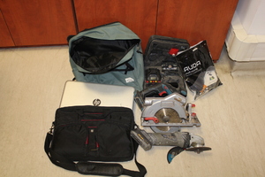 Na zdjęciu znajduje się niebieski plecak, laptop, czarny plecak, szlifierka w kolorze srebrnym oraz czarna walizka z zawartością narzędzi.